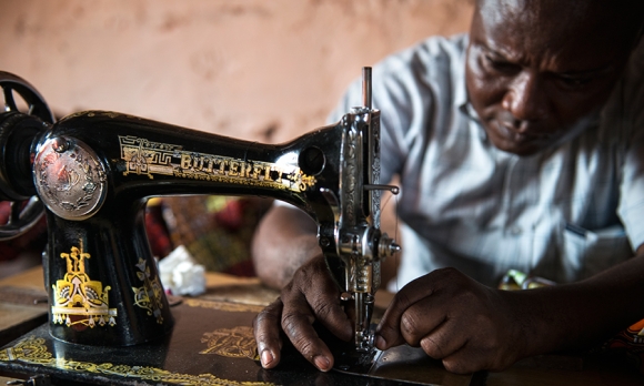 Gaby Ngabu Kasongo sewing a dress. The effects of Konzo make it difficult for Gaby to work long hours. He says his hands begin to get stiff and hurt when working. He also struggles to use his legs to operate the sewing machine. 