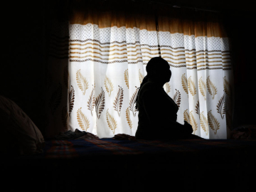 A woman, shown in shadow, sits posing in profile against the backdrop of a light white curtain with beige and brown designs.