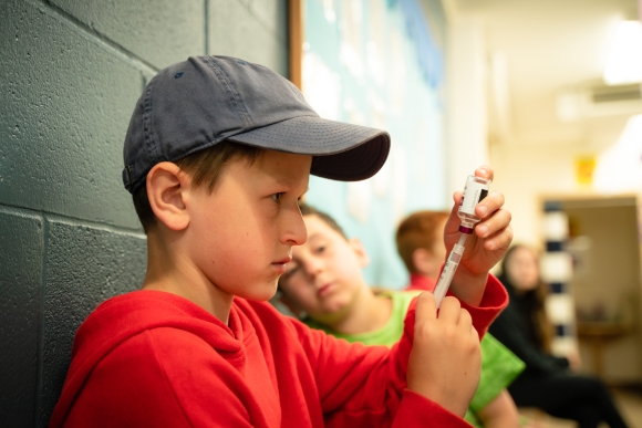 A young boy in a grey baseball cap and red hoodie preps a syringe with insulin 