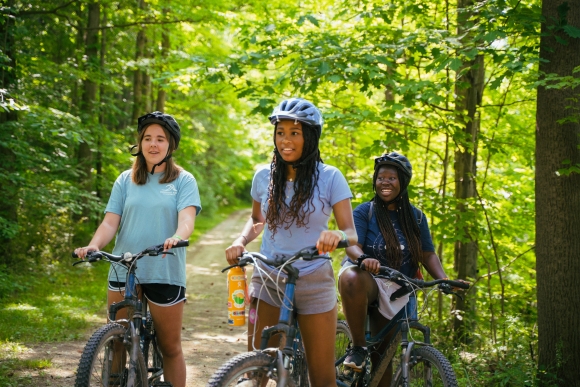 3 teen girls on bikes pause on a path in the woods at Camp Ho Mita Koda